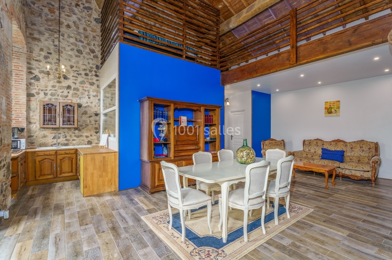 Salle à manger avec table blanche, chaises, murs en pierre et bleu, plafond en bois et coin salon en arrière-plan.