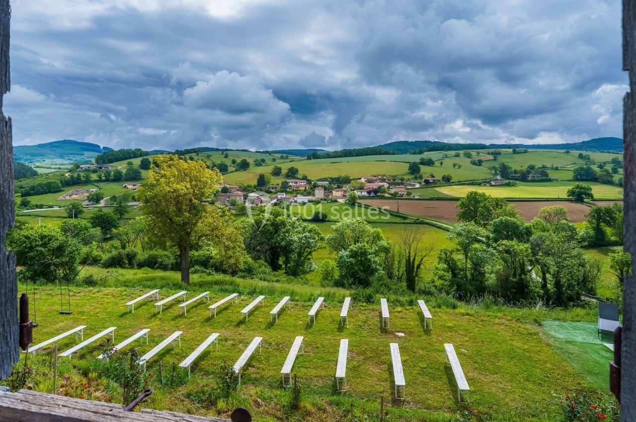 Vue sur un paysage rural avec des bancs alignés en plein air, entourés de champs verdoyants et d'un village au loin.