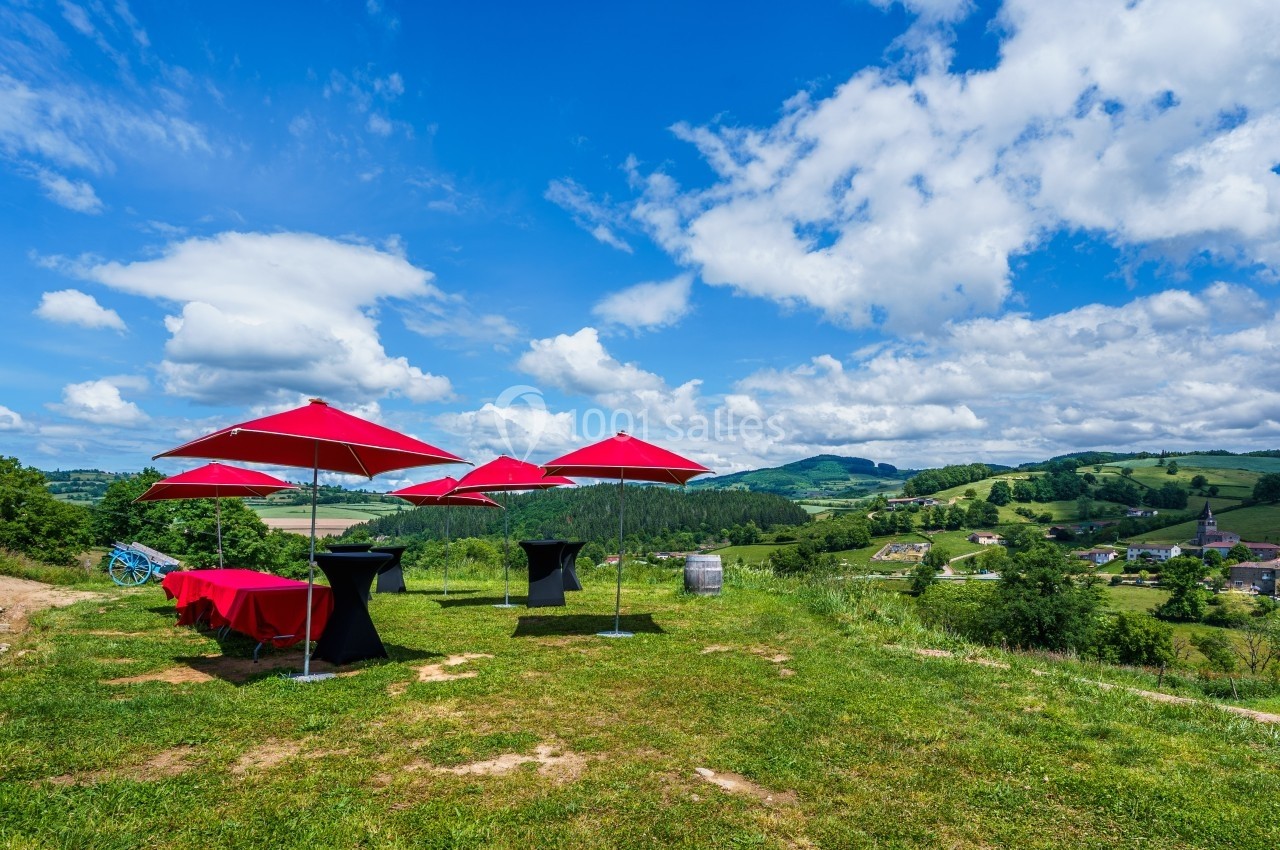 Vue sur une colline verdoyante avec des parasols rouges et des tables noires, surplombant un village et des champs.