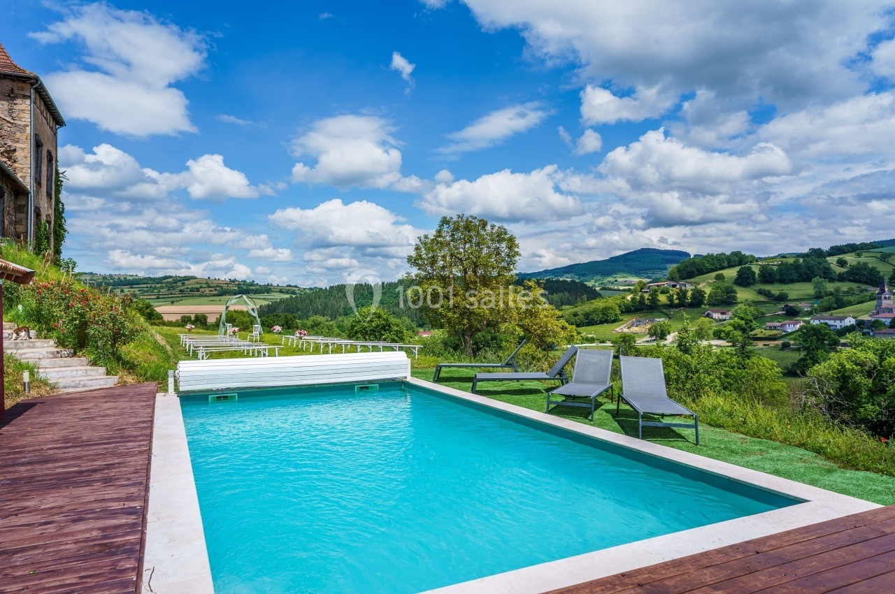 Piscine extérieure entourée de chaises longues, avec vue sur une campagne vallonnée sous un ciel partiellement nuageux.