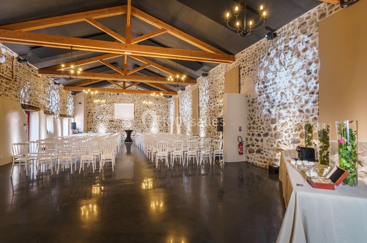 Salle spacieuse en pierre avec plafond en poutres apparentes, rangées de chaises blanches et table décorative à droite.