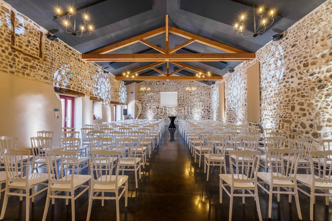 Salle de réception en pierre avec des chaises blanches alignées, un plafond en poutres apparentes et des lustres suspendus.