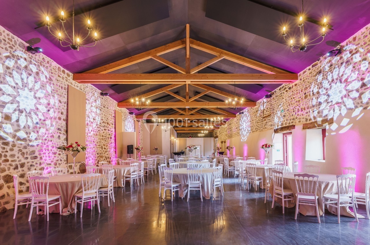 Salle de réception décorée avec des tables rondes, des chaises blanches et un éclairage rose sur des murs en pierre.