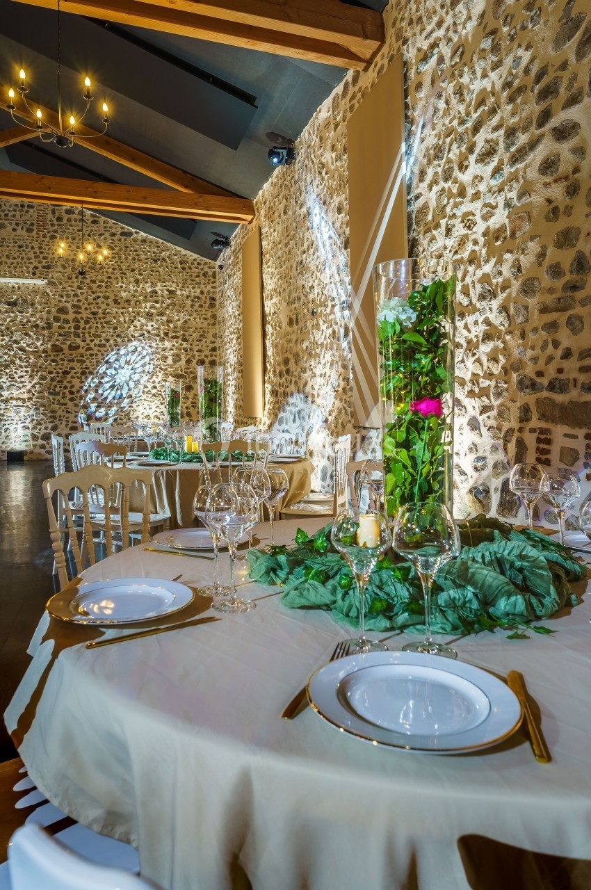 Salle de réception en pierre avec tables décorées de nappes blanches, vaisselle élégante et centres de table végétaux.