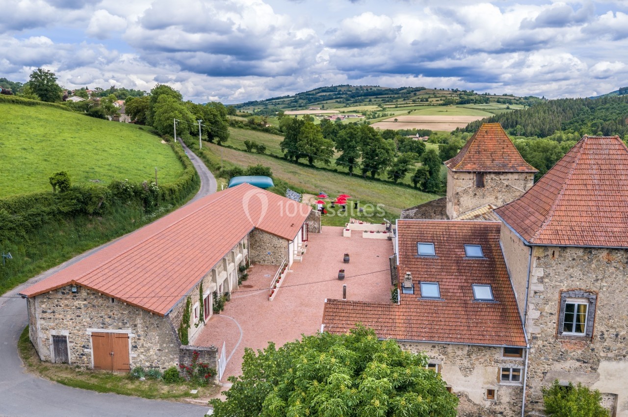 Vue aérienne d'un bâtiment en pierre avec une cour pavée, entouré de collines verdoyantes sous un ciel nuageux.