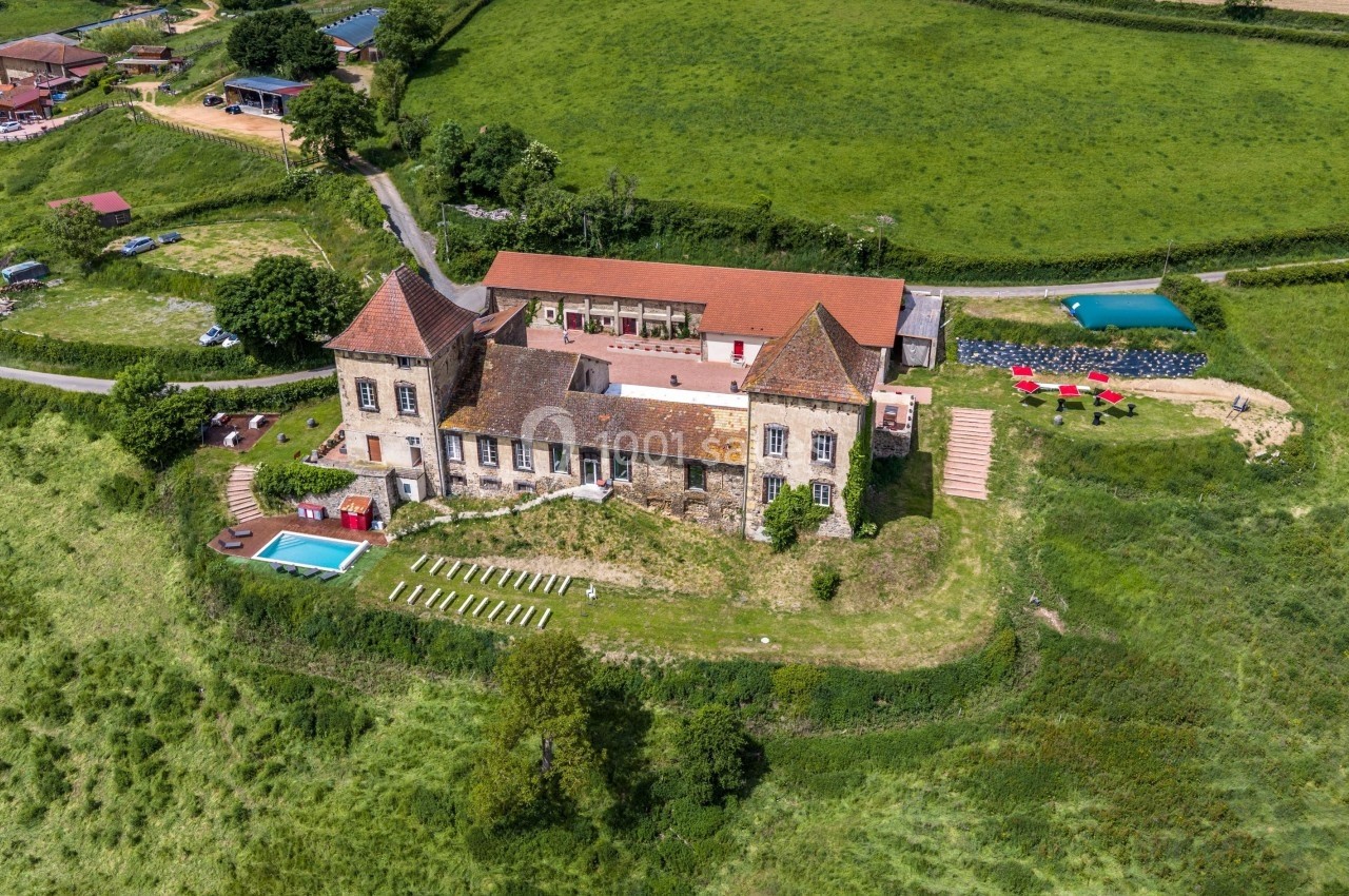Vue aérienne d'un château en pierre avec piscine, jardin aménagé et bâtiments annexes, entouré de champs verdoyants.