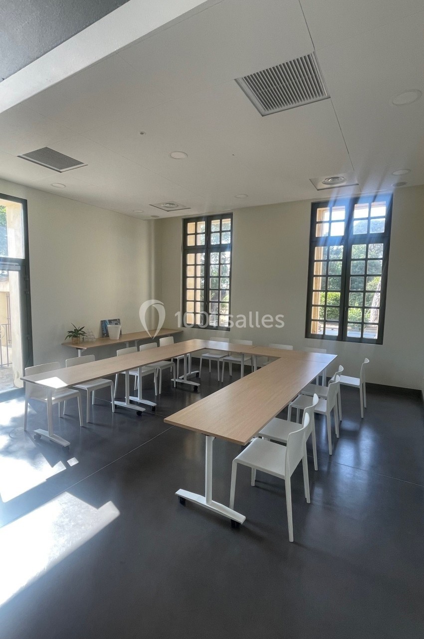 Salle lumineuse avec de grandes fenêtres, tables en U et chaises blanches, sol sombre et murs clairs.