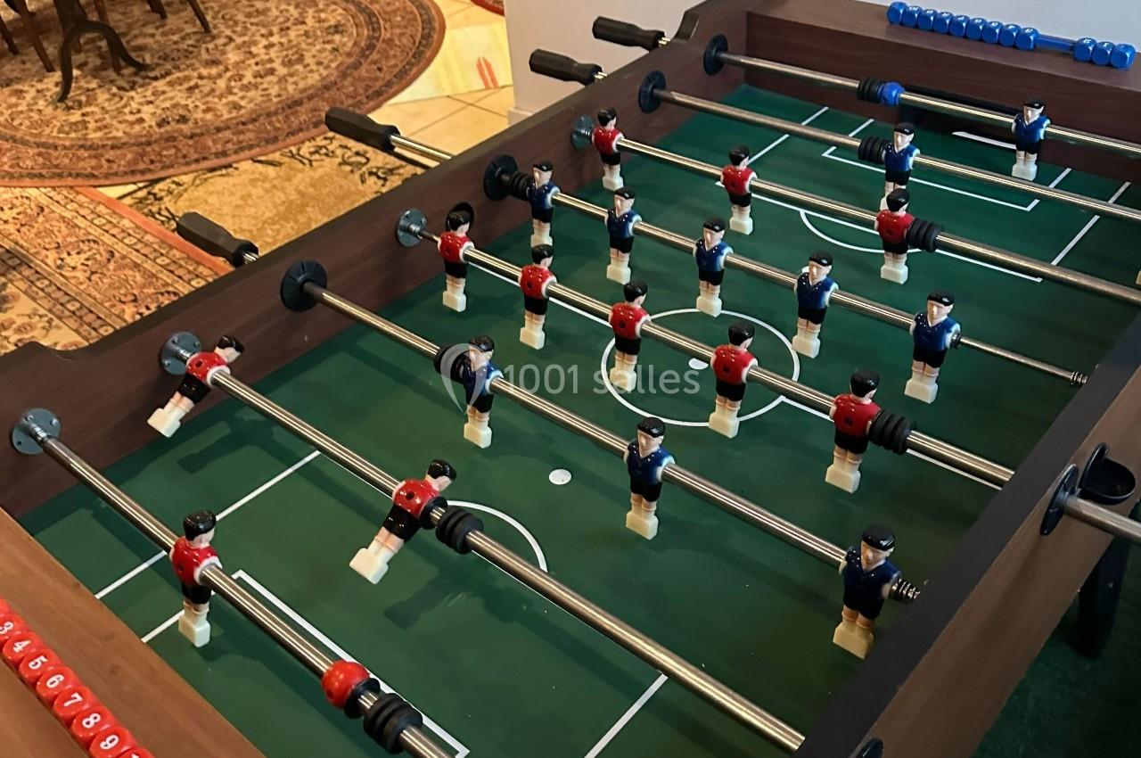 Table de baby-foot avec figurines rouges et bleues alignées sur un terrain vert, dans un environnement intérieur.