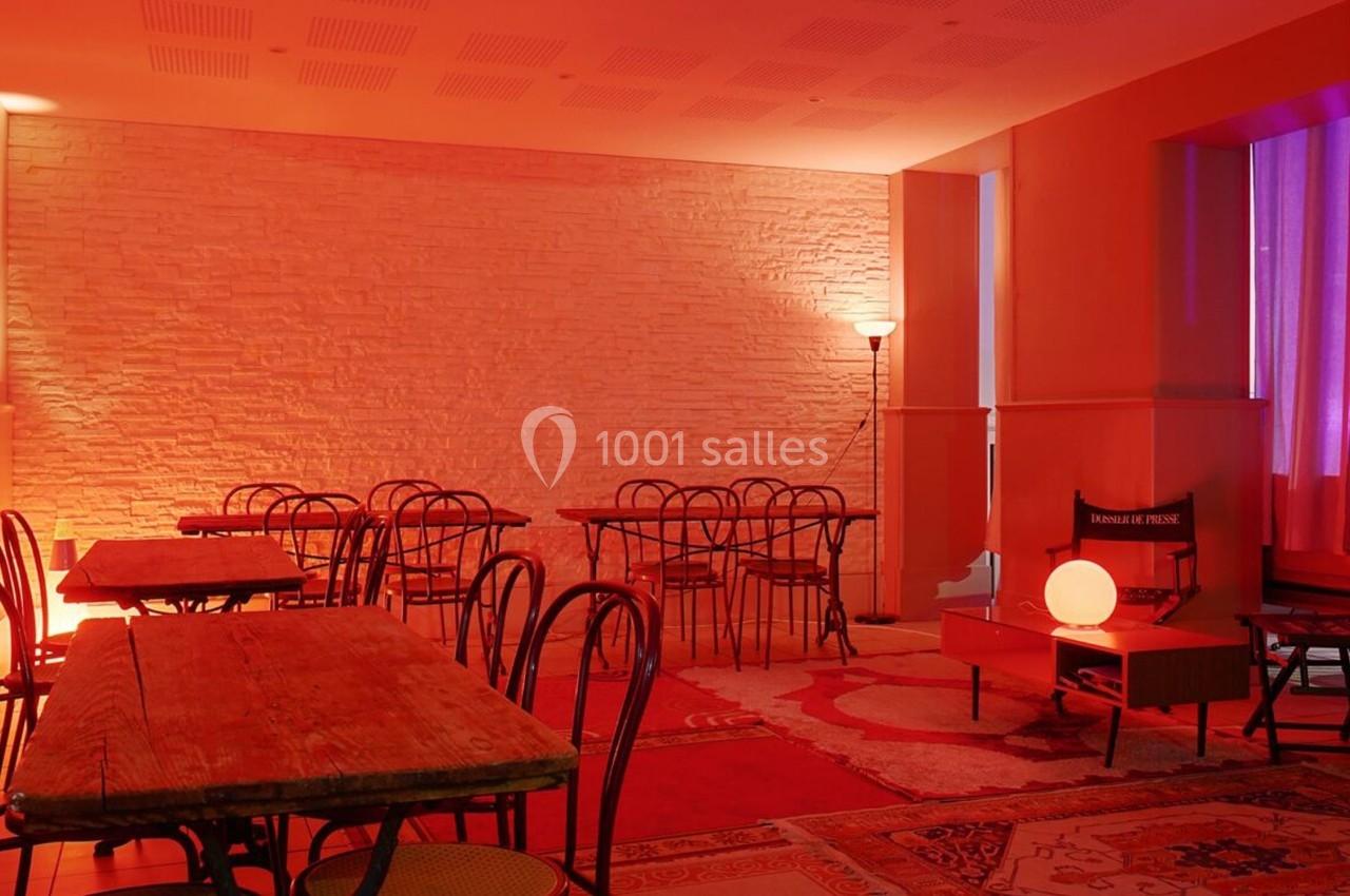 Salle avec tables en bois, chaises noires et éclairage rouge tamisé, murs en briques et tapis au sol.