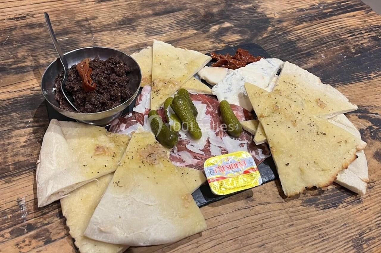 Assiette de charcuterie avec pain plat, cornichons, tapenade, fromage et beurre sur une table en bois.