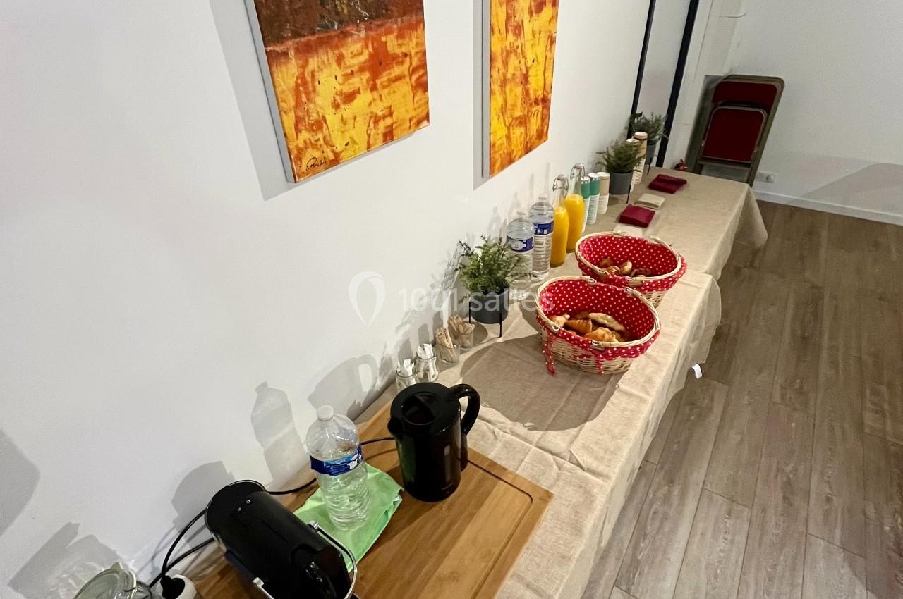 Table avec cafetière, carafe, paniers de viennoiseries, jus de fruits et décorations murales dans une pièce lumineuse.