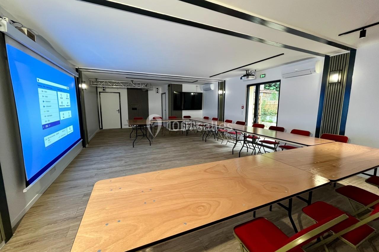 Salle de réunion moderne avec tables en bois clair disposées en U, chaises rouges et écran de projection allumé.