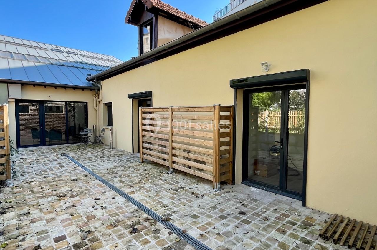 Cour pavée avec façade beige, grandes baies vitrées, clôtures en bois et ciel dégagé en arrière-plan.
