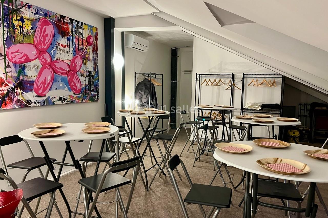 Salle mansardée aménagée avec des tables rondes, des chaises pliantes noires et un décor mural coloré.