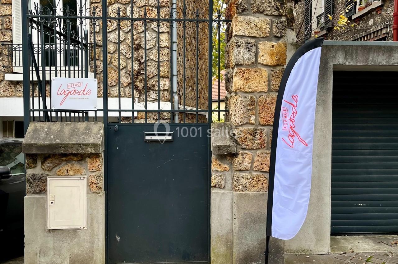 Portail noir en fer forgé encadré par un mur en pierre, avec une enseigne et un drapeau publicitaire à droite.