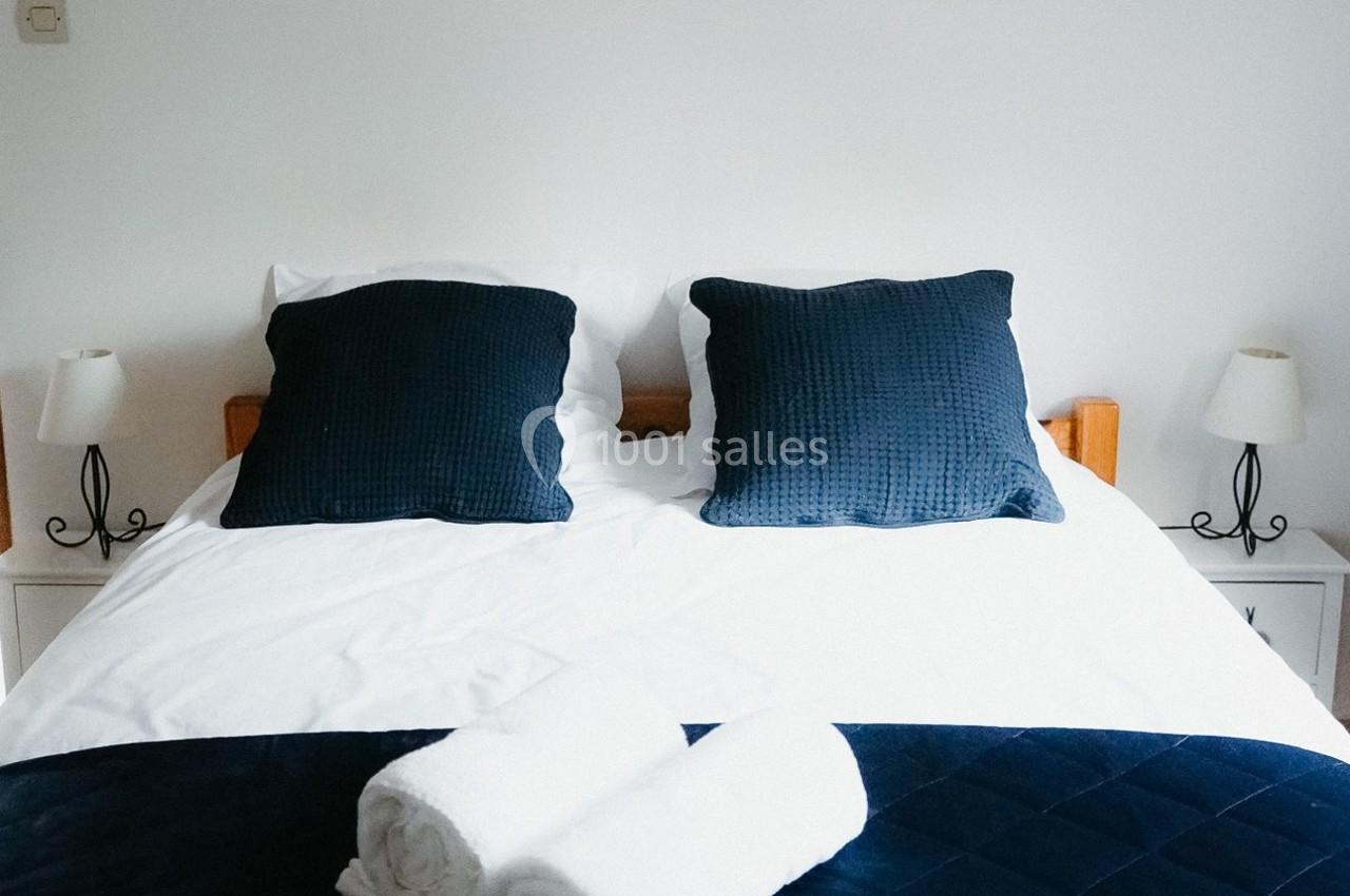 Lit double avec draps blancs, coussins bleus, couverture assortie et deux serviettes roulées, encadré par des lampes.