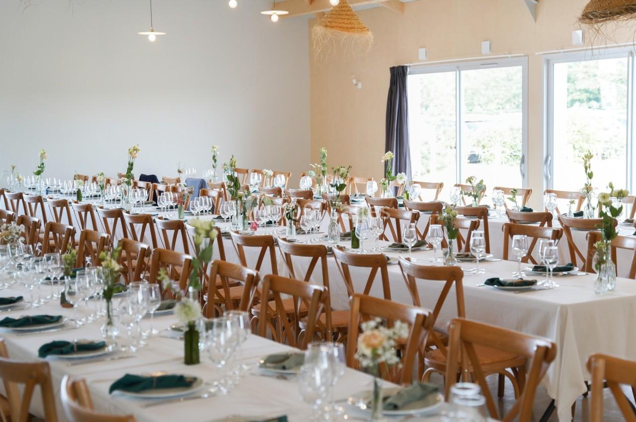 Salle de réception lumineuse avec de longues tables dressées, décorées de fleurs et de serviettes vertes.