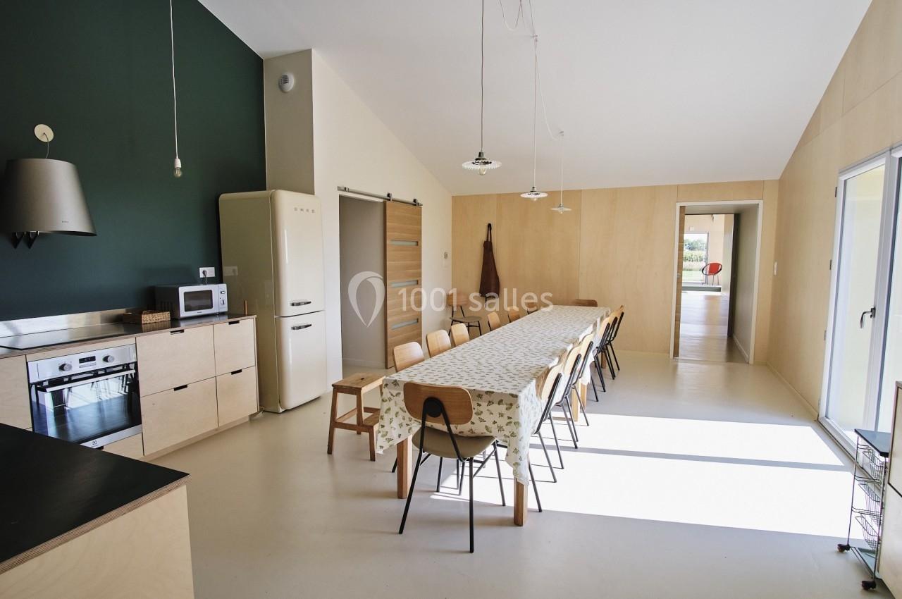 Cuisine moderne avec table longue, chaises en bois, électroménagers et lumière naturelle entrant par une baie vitrée.