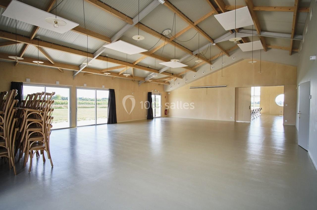 Salle spacieuse avec sol clair, plafond en bois, grandes fenêtres et chaises empilées dans un coin.