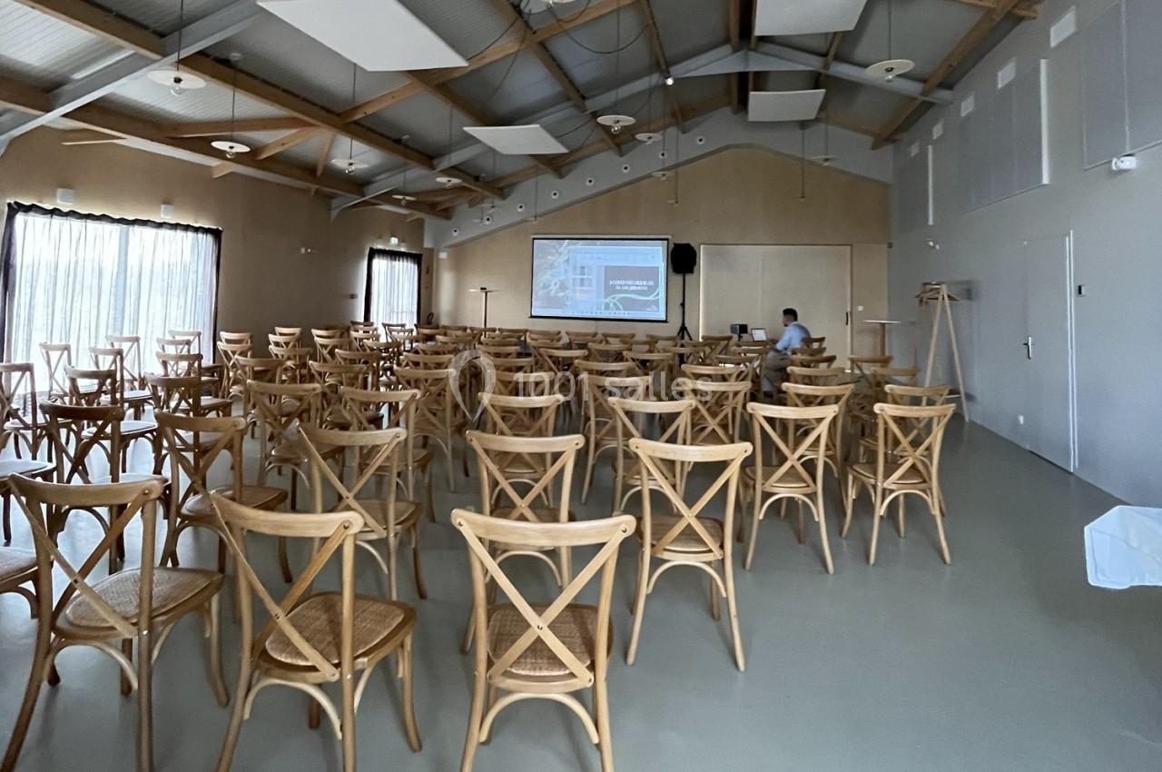 Salle de conférence lumineuse avec des rangées de chaises en bois et un écran de projection allumé au fond.