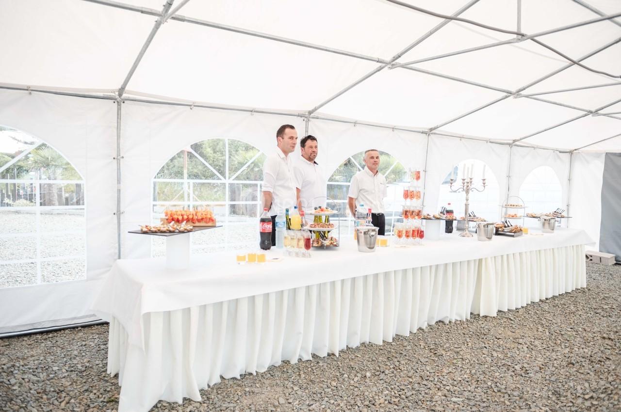 Trois serveurs derrière une table garnie de boissons et amuse-bouches sous une tente blanche.