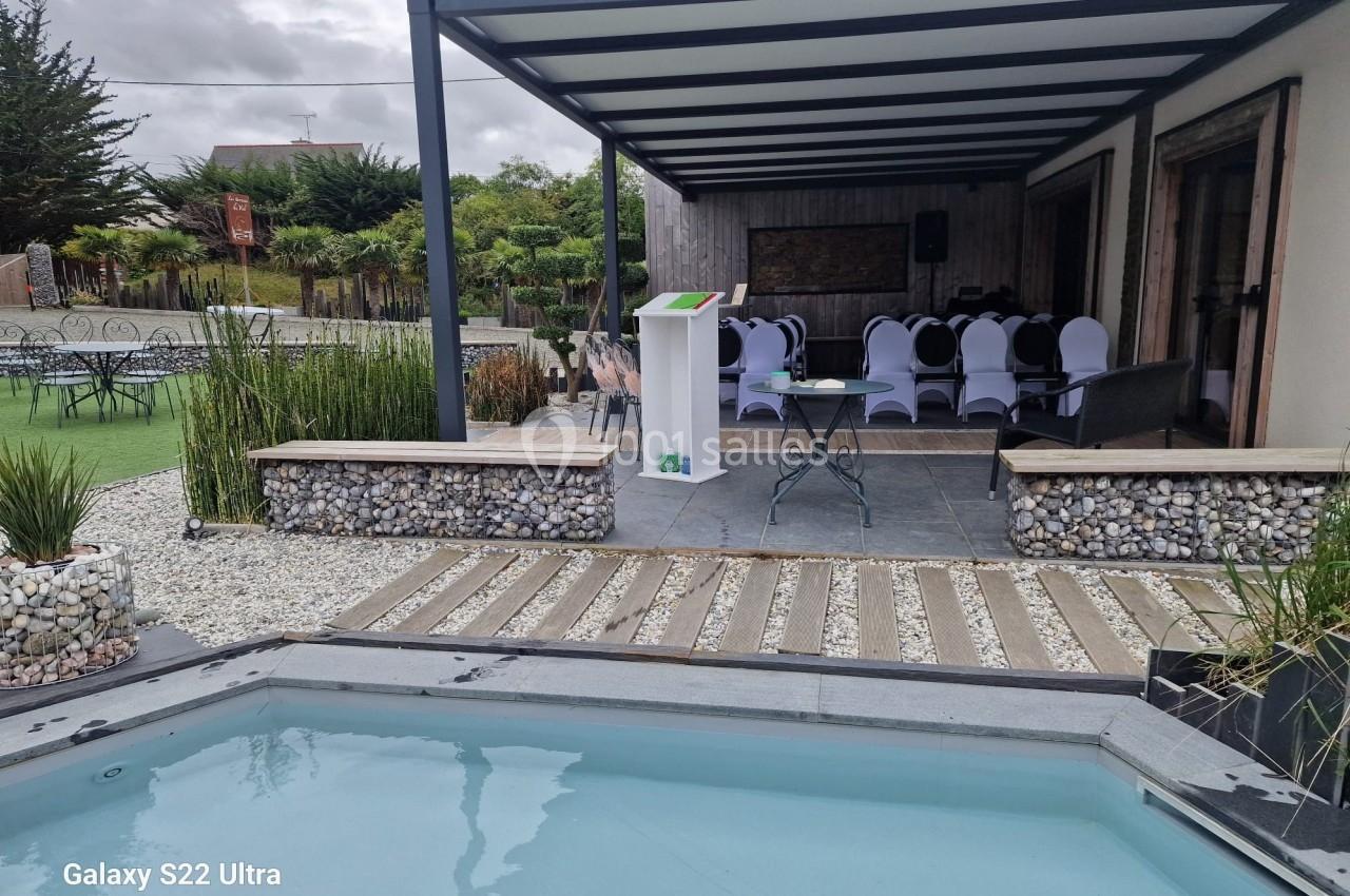 Terrasse couverte aménagée avec des chaises blanches, un pupitre, et une piscine au premier plan dans un jardin paysager.