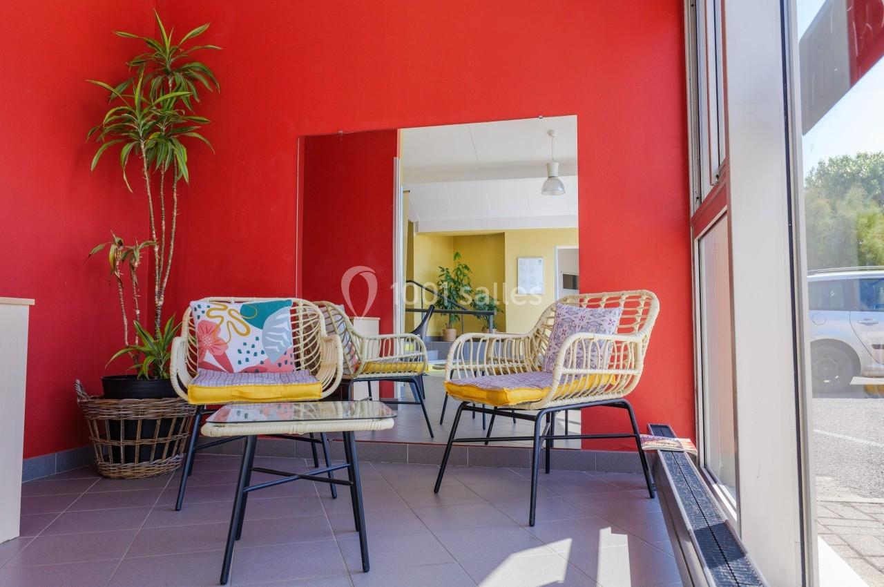Deux fauteuils en rotin avec coussins colorés et une table basse dans une pièce aux murs rouges, près d'une baie vitrée.