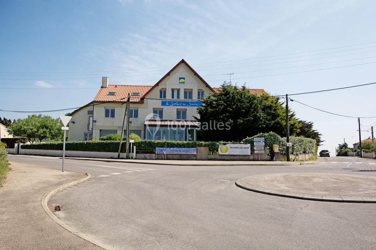 Bâtiment à deux étages avec toit en tuiles rouges, situé à une intersection bordée de végétation et de panneaux.