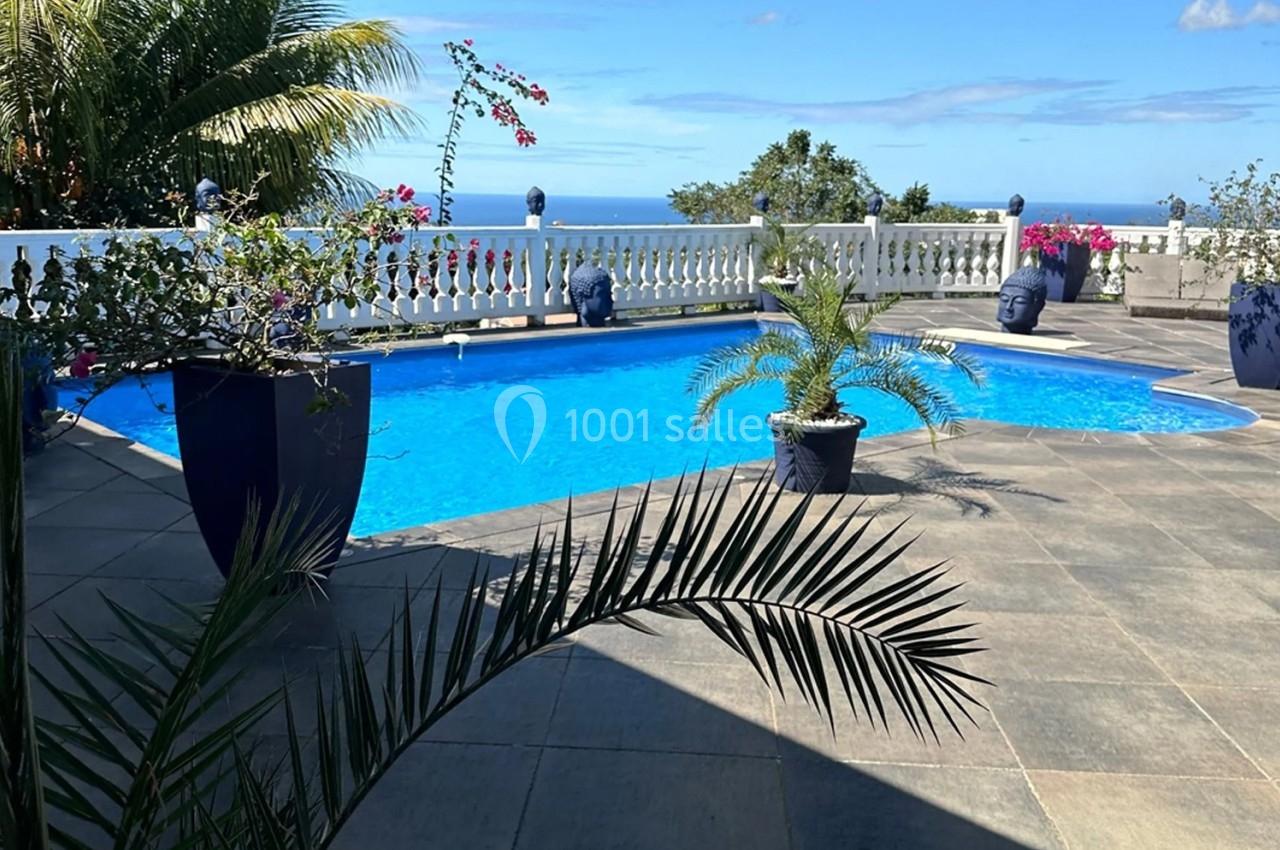 Piscine extérieure entourée de plantes, terrasse carrelée et vue dégagée sur l'océan sous un ciel bleu.