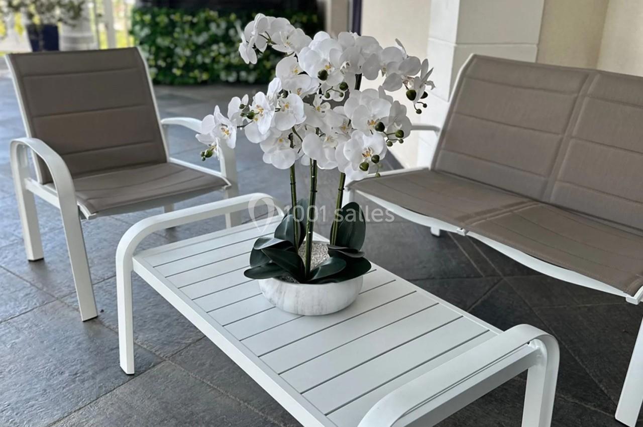Orchidée blanche dans un pot en marbre sur une table basse blanche entourée de fauteuils sur une terrasse carrelée.