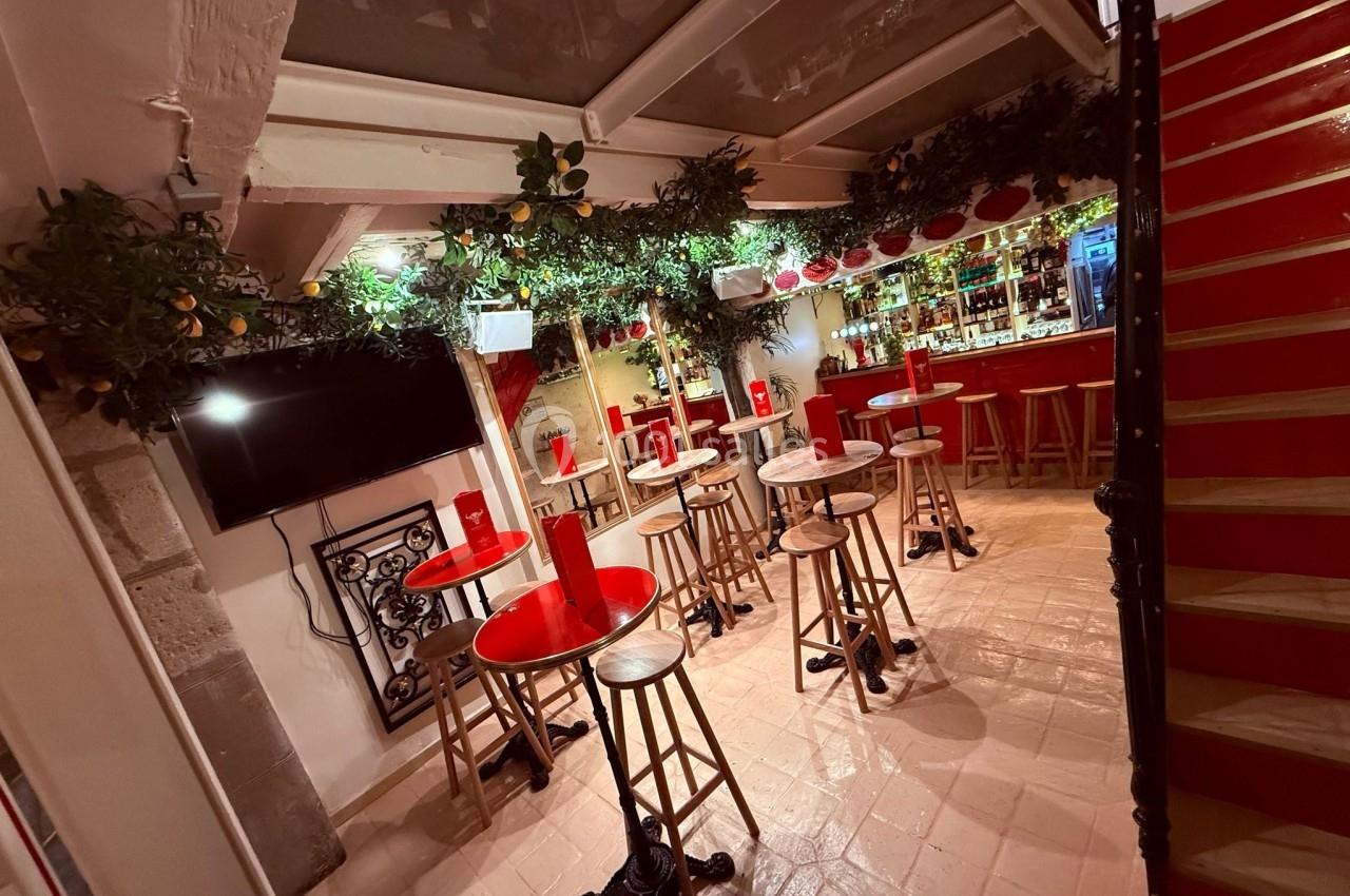 Salle de bar avec tables hautes, tabourets en bois, décorations végétales et comptoir éclairé en arrière-plan.