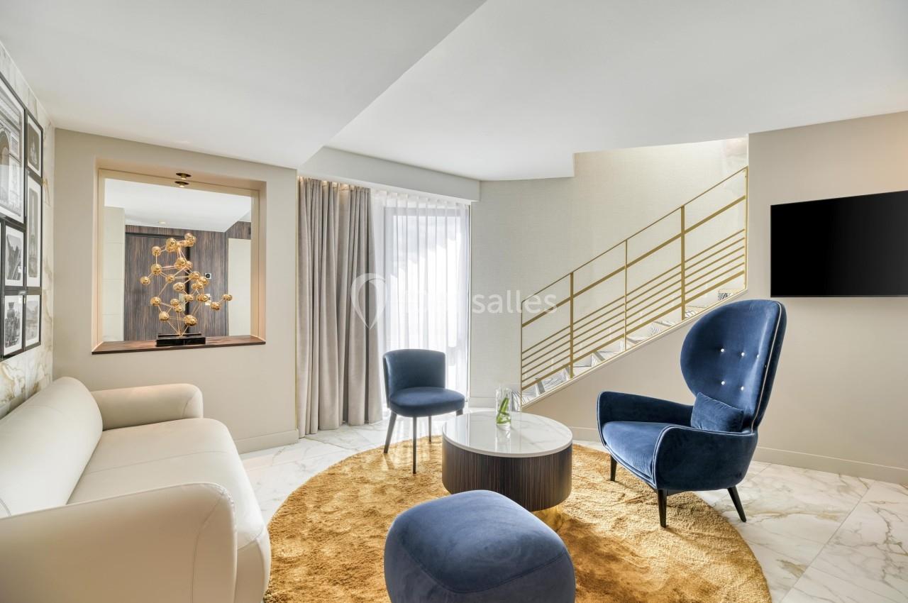Salon moderne avec canapé blanc, fauteuils bleus, table basse ronde, tapis beige et escalier en arrière-plan.