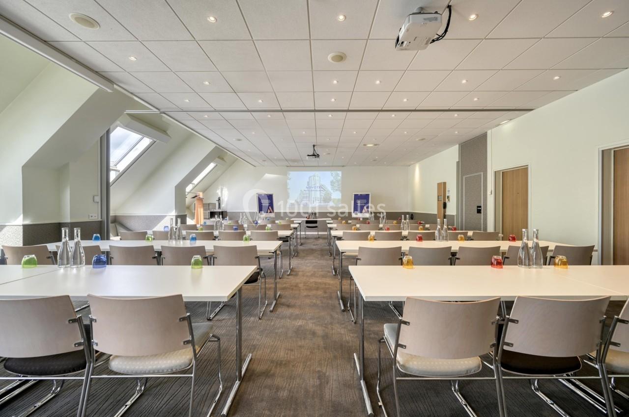 Salle de conférence lumineuse avec des rangées de tables et chaises, équipée d'un projecteur et d'une présentation affichée.