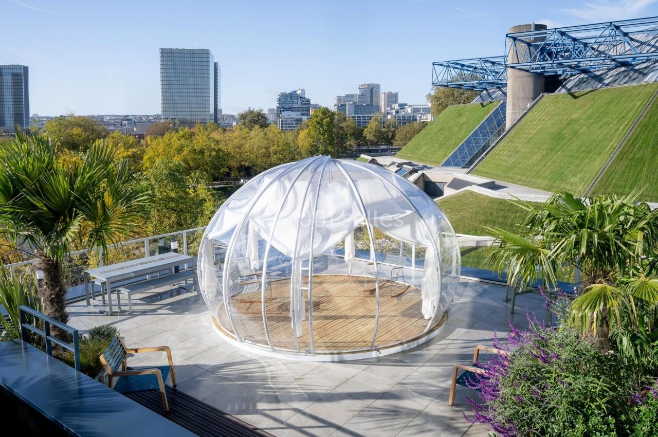 Dôme transparent installé sur une terrasse entourée de végétation, avec vue sur des bâtiments urbains modernes.