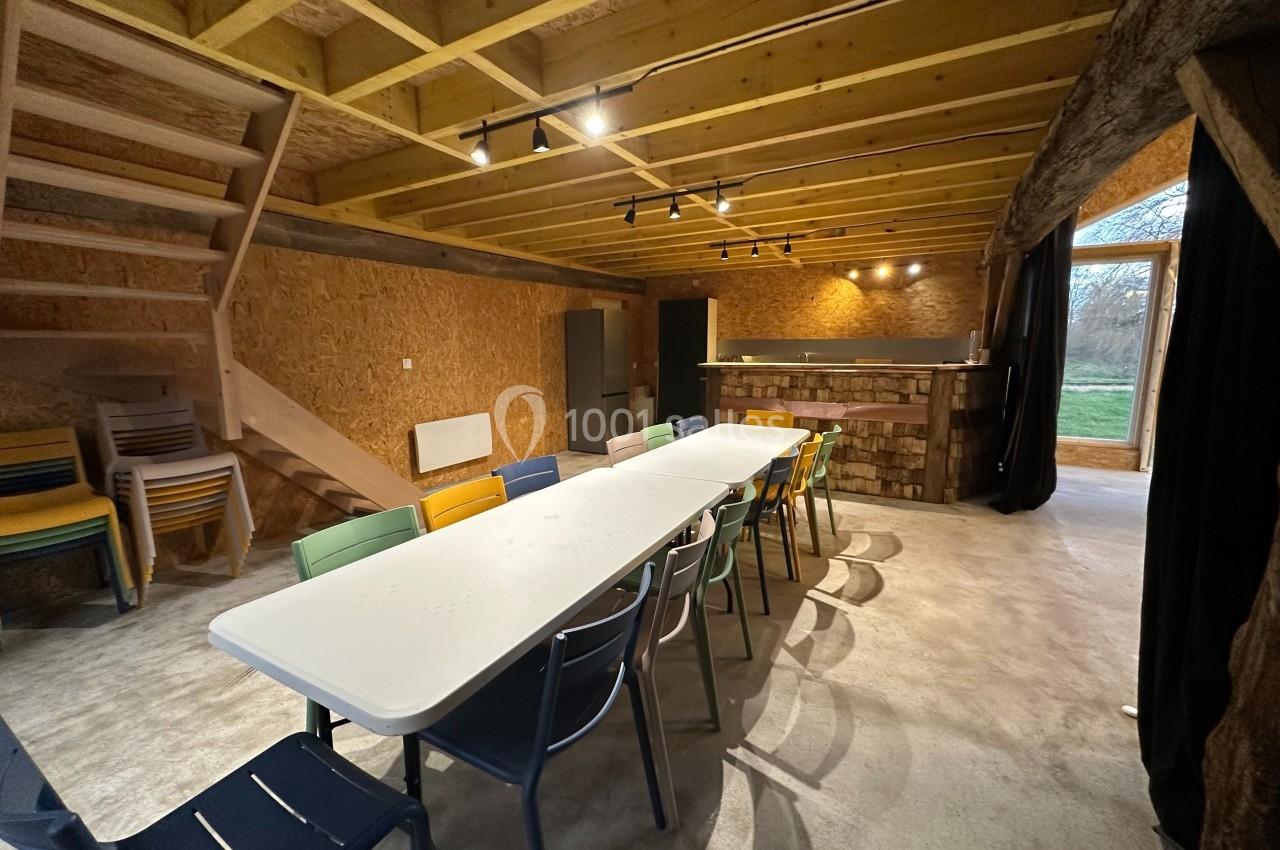 Salle avec une grande table entourée de chaises colorées, éclairée par des spots, avec un escalier et un bar en bois.