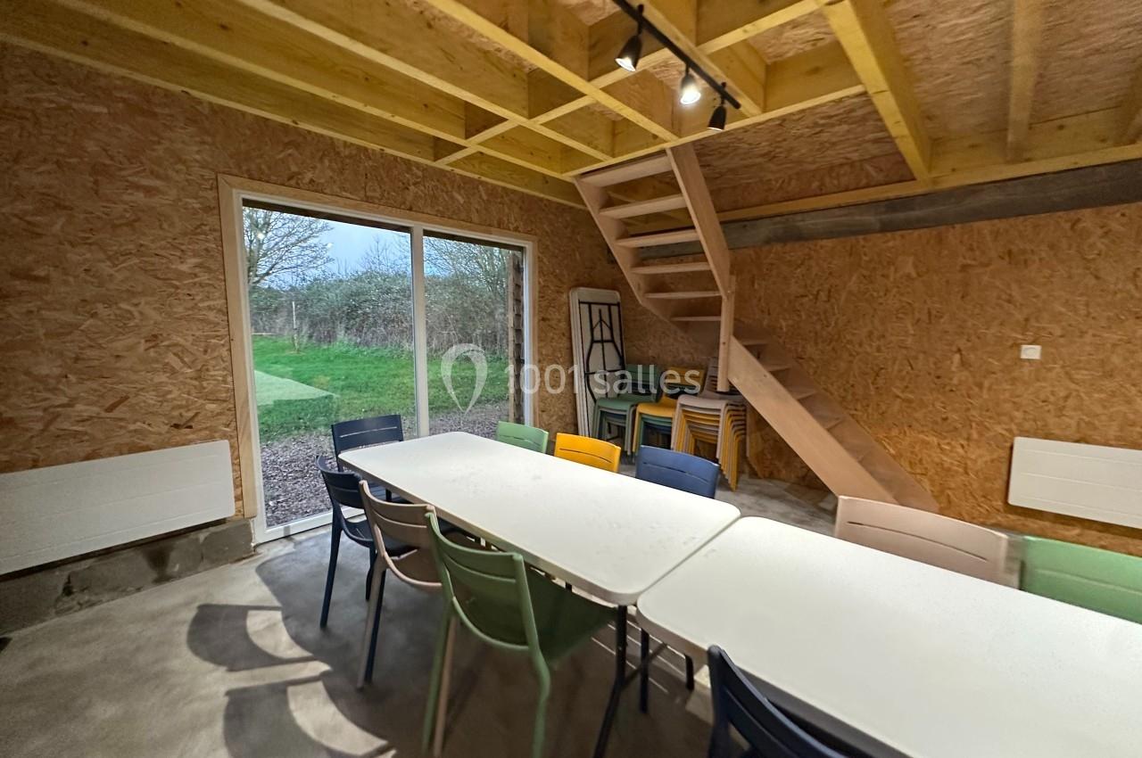 Salle en bois avec une grande table entourée de chaises colorées, escalier en bois et baie vitrée donnant sur un jardin.