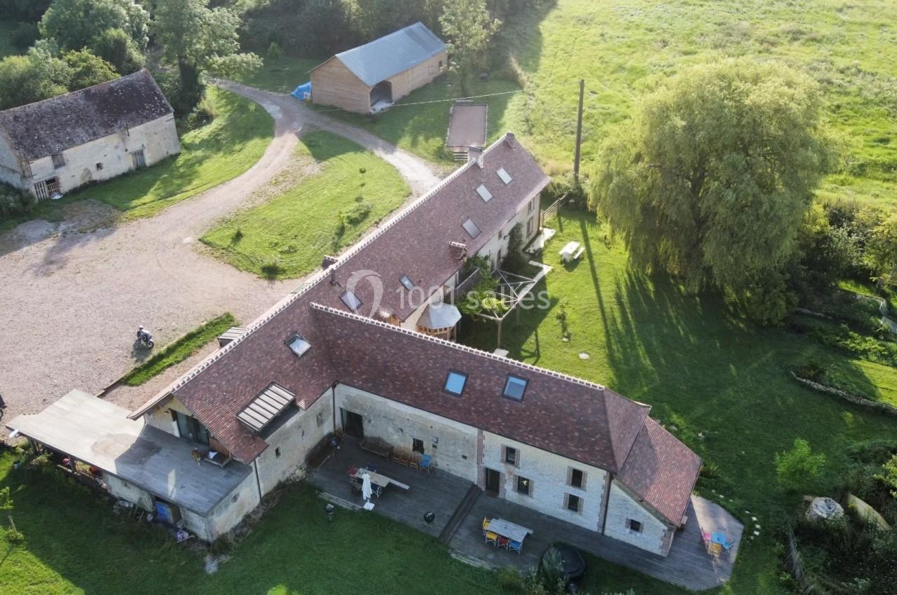 Vue aérienne d'une ferme en pierre avec toits en tuiles rouges, entourée de verdure et de bâtiments annexes.