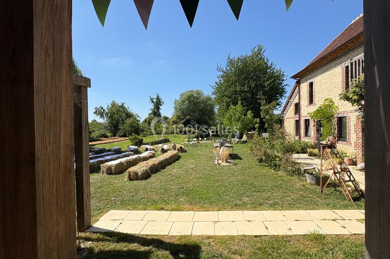 Vue depuis un cadre en bois sur un jardin ensoleillé avec des bottes de paille, des guirlandes et une maison en pierre.