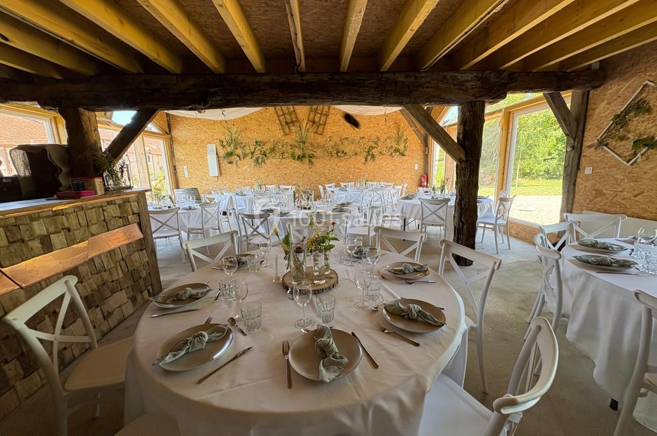 Salle de réception rustique avec tables rondes dressées, décorations végétales et poutres apparentes en bois.