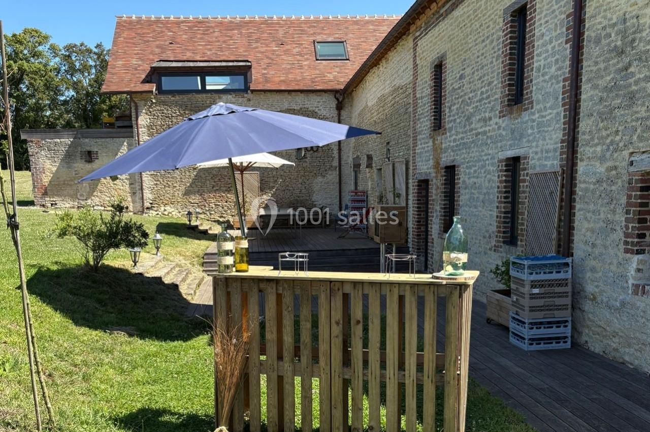 Cour extérieure avec terrasse en bois, parasol bleu, table en bois et bâtiments en pierre sous un ciel dégagé.