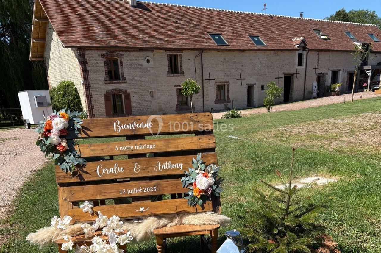 Panneau en bois décoré de fleurs, annonçant un mariage, devant une bâtisse en pierre avec un toit en tuiles.