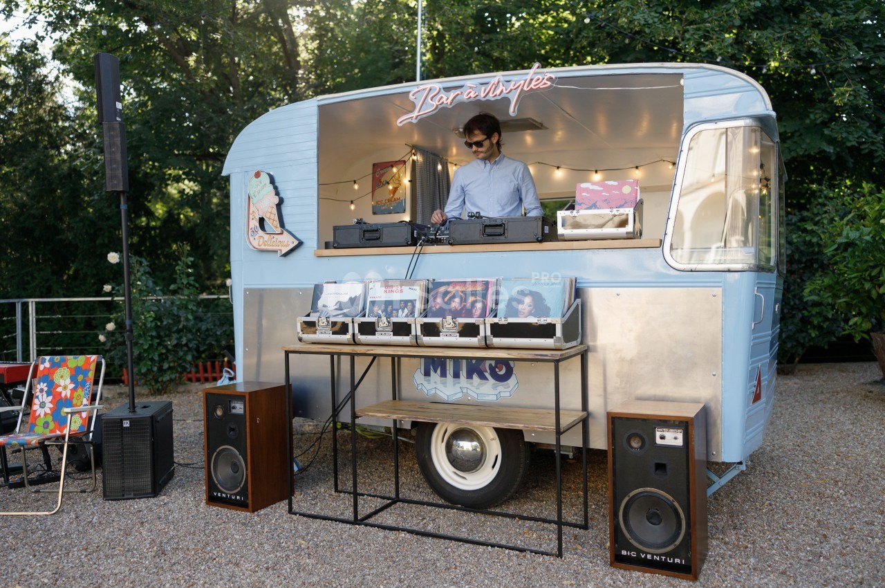 Un DJ mixe de la musique dans une caravane aménagée, entourée de vinyles et d'enceintes, en extérieur.