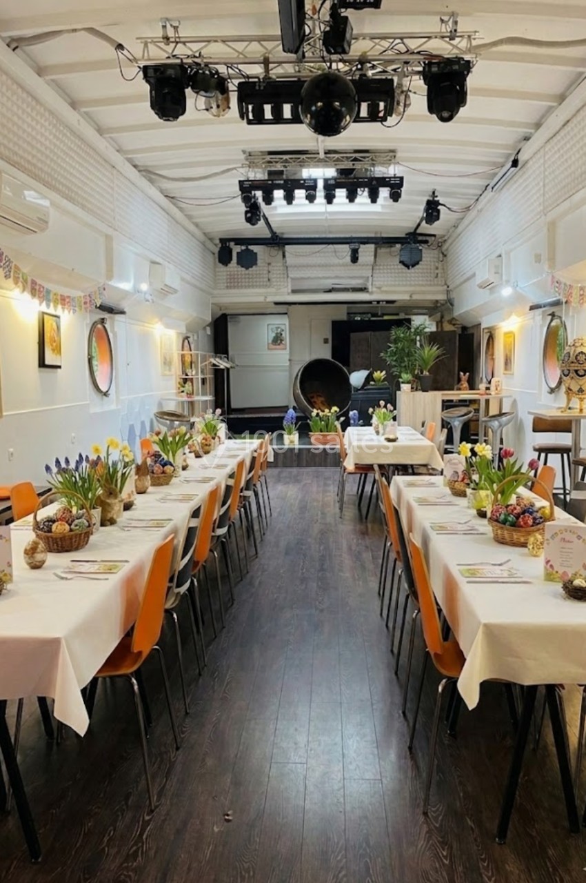 Salle décorée pour un événement, avec deux longues tables dressées, des fleurs et des guirlandes colorées.