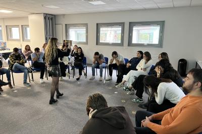 Quatre personnes jouent à un jeu de cartes autour d'une table dans une salle lumineuse avec d'autres personnes en arrière…