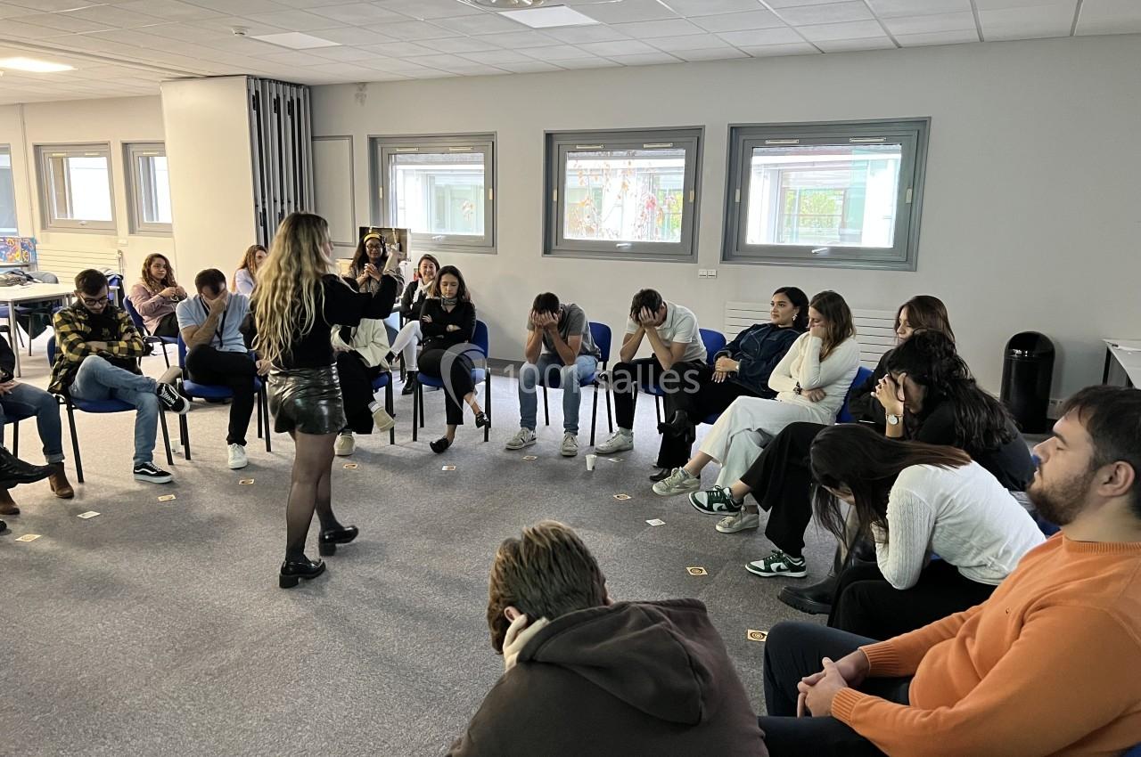 Un groupe de personnes assises en cercle dans une salle, écoutant une femme debout qui s'exprime.