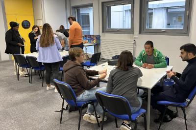 Quatre personnes jouent à un jeu de cartes autour d'une table dans une salle lumineuse avec d'autres personnes en arrière…