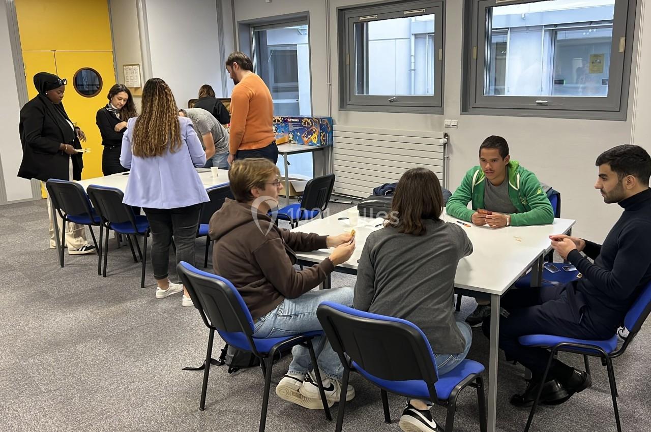 Des personnes discutent et travaillent en groupe autour de tables dans une salle lumineuse avec des fenêtres.