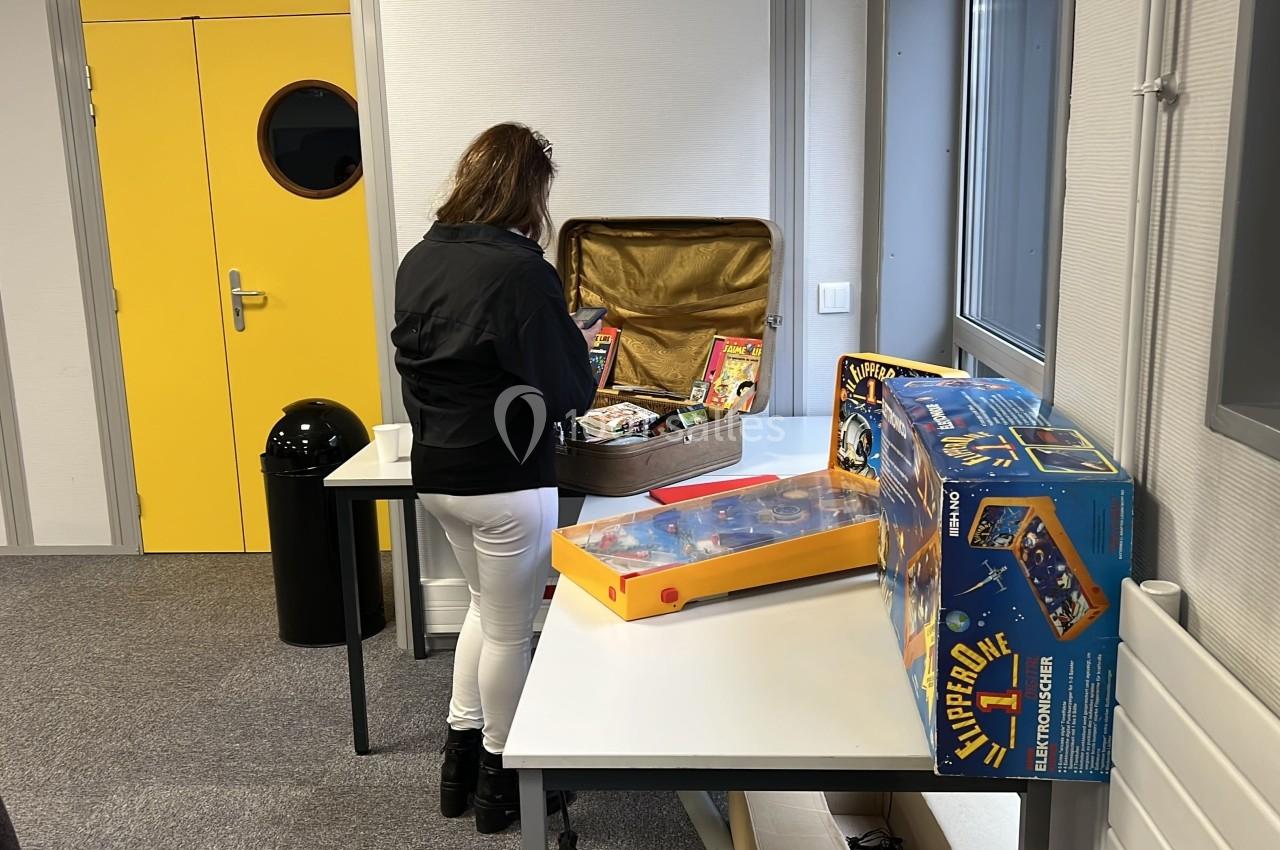 Une femme examine des objets dans une valise ouverte sur une table, entourée de jeux rétro dans une pièce lumineuse.