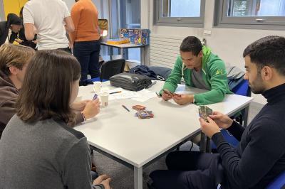 Quatre personnes jouent à un jeu de cartes autour d'une table dans une salle lumineuse avec d'autres personnes en arrière…