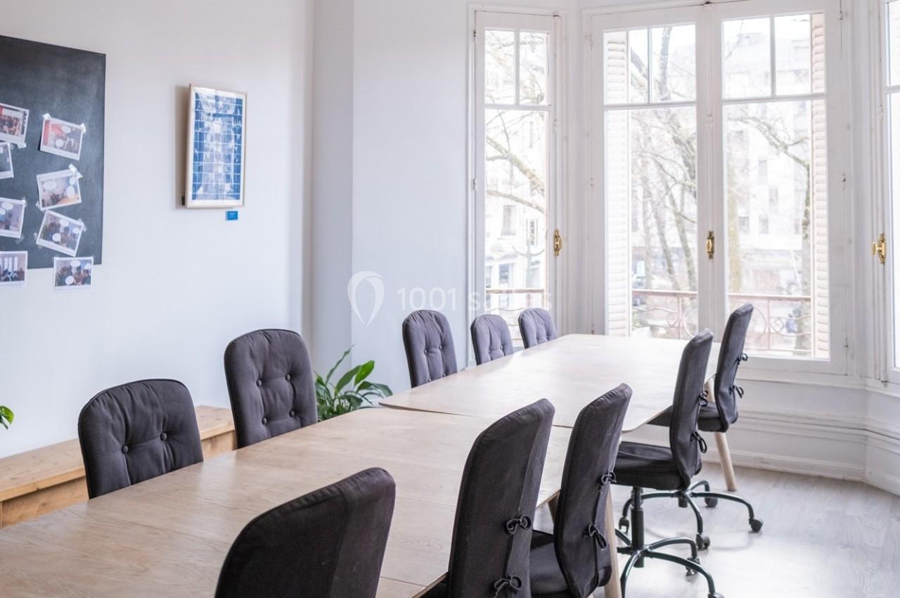 Salle de réunion lumineuse avec une grande table en bois entourée de chaises, près de fenêtres donnant sur l'extérieur.